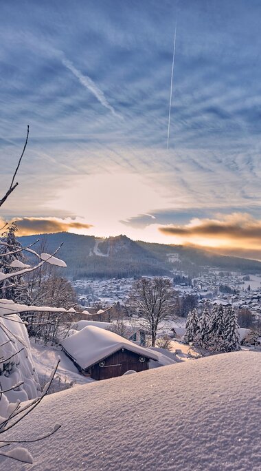 Über glitzernden Schnee hinweg blickt man auf Bodenmais. Über der Ortschaft wacht der Silberberg, hinter dem die Sonne hervor blitzt, der blaue Himmel ist durchzogen von Schleierwolken. | © Bodenmais Tourismus & Marketing GmbH