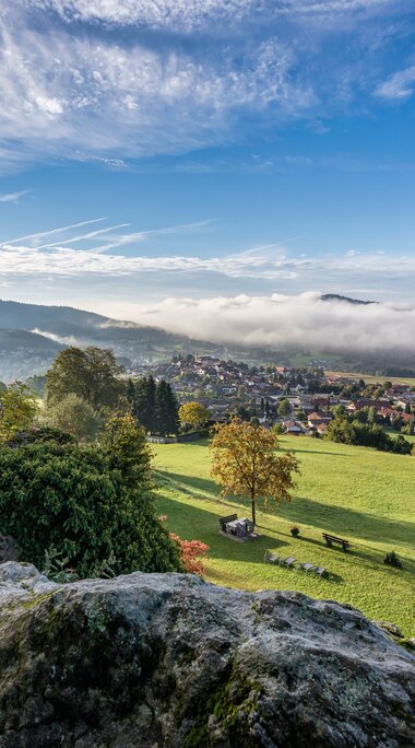 Vom Wanderparkplatz Klause aus blickt man hinab auf Bodenmais. Grüne Wiesen leuchten in der Sonne. Im Tal hängt teilweise Nebel. | © Bodenmais Tourismus & Marketing GmbH