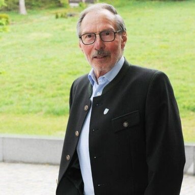 Ein älterer Herr mit Brille in einem blauen Hemd mit dunklem Sakko steht vor einer Grünfläche und blickt freundlich in die Kamera.  | © Markt Bodenmais