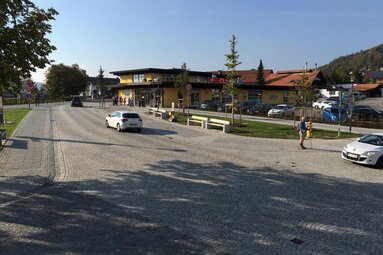 Auf einer Kreuzung sieht man drei Fahrzeuge. Dahinter Geschäfte und einen Parkplatz. Ein großer Baum wirft einen Schatten auf die Straße.  | © Markt Bodenmais