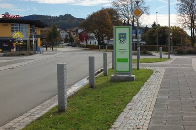 Neben der Straße sieht man einen Grünstreifen mit Gehweg. Im Hintergrund eine Landschaft mit Häusern und Bäumen. Ein Schild zeigt das Wappen von Bodenmais.  | © Markt Bodenmais