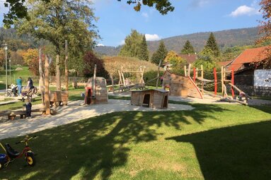 Das Bild zeigt einen Spielplatz mit Holzspielgeräten. Der Untergrund des Spielplatzes besteht aus Sand. Rings um den Sand ist Wiese. Im Hintergrund erhebt sich ein Waldgebiet.  | © Markt Bodenmais