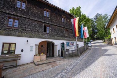 An einer Kopfsteinstraße steht ein historisch anmutendes, dreistöckiges Haus mit Holzschindeln an der Fassade. | © Bodenmais Tourismus & Marketing GmbH